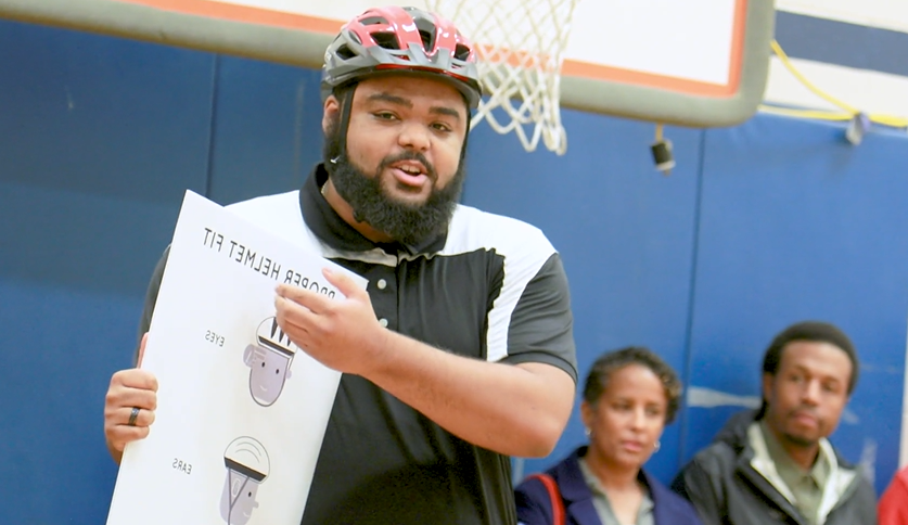 PE teacher speaking 和 holding sign that says "proper helmet fit."